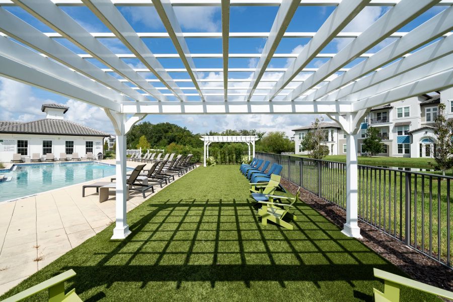 Shadetree Apartments with White pergola over green lawn with lounge chairs by a pool, with buildings and trees in the background.