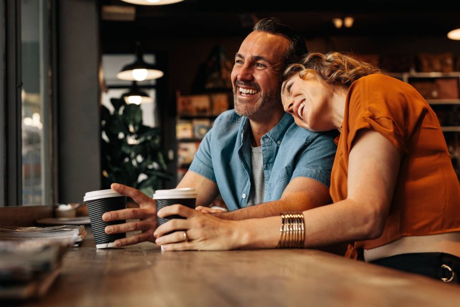 Shadetree Apartments with a couple smiling and the woman putting her head on the shoulder of man while sitting at a coffee shop. Smiling couple at a café.