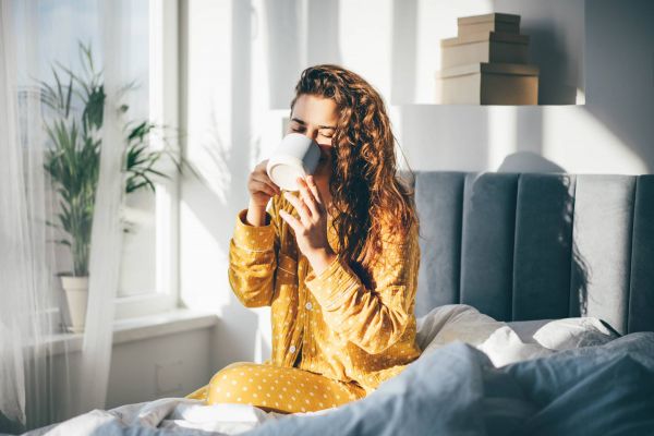 Shadetree Apartments with Woman in yellow pajamas sits on a bed by a window, enjoying a hot drink from a white mug in the morning light.