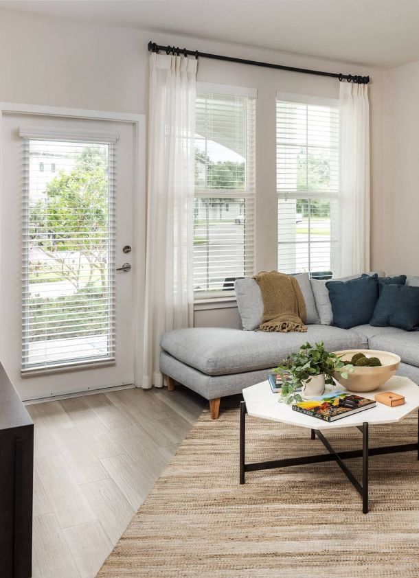 Shadetree Apartments with Bright living room with a gray sectional sofa, coffee table, and large windows with white curtains and blinds.