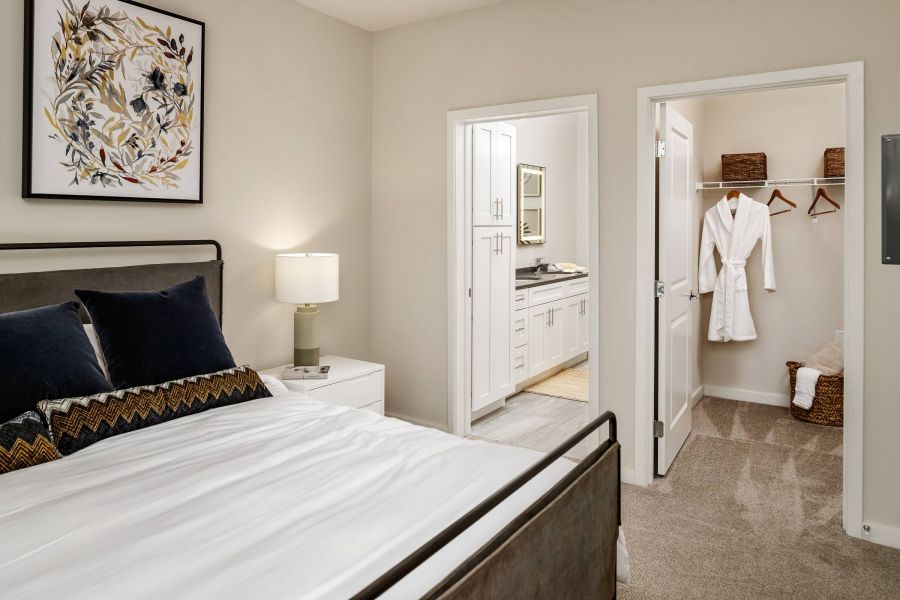Shadetree Apartments with Modern bedroom with neatly made bed, nightstand, art, and view into bathroom and walk-in closet with robes.